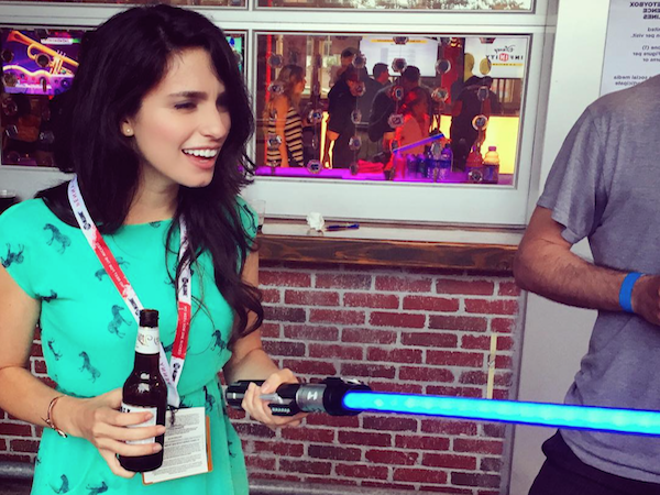 Pretty brunette with a beer in one hand and a light saber in other poses in blue dress