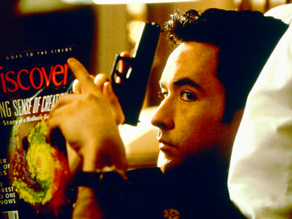 John Cusack in a black tee holds a gun while reading Discover magazine!